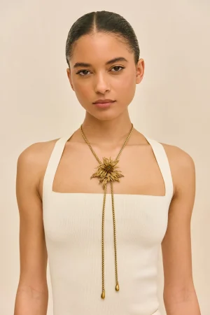 Woman wearing a white sleeveless top with a gold sunburst pendant necklace, facing the camera against a beige background.
