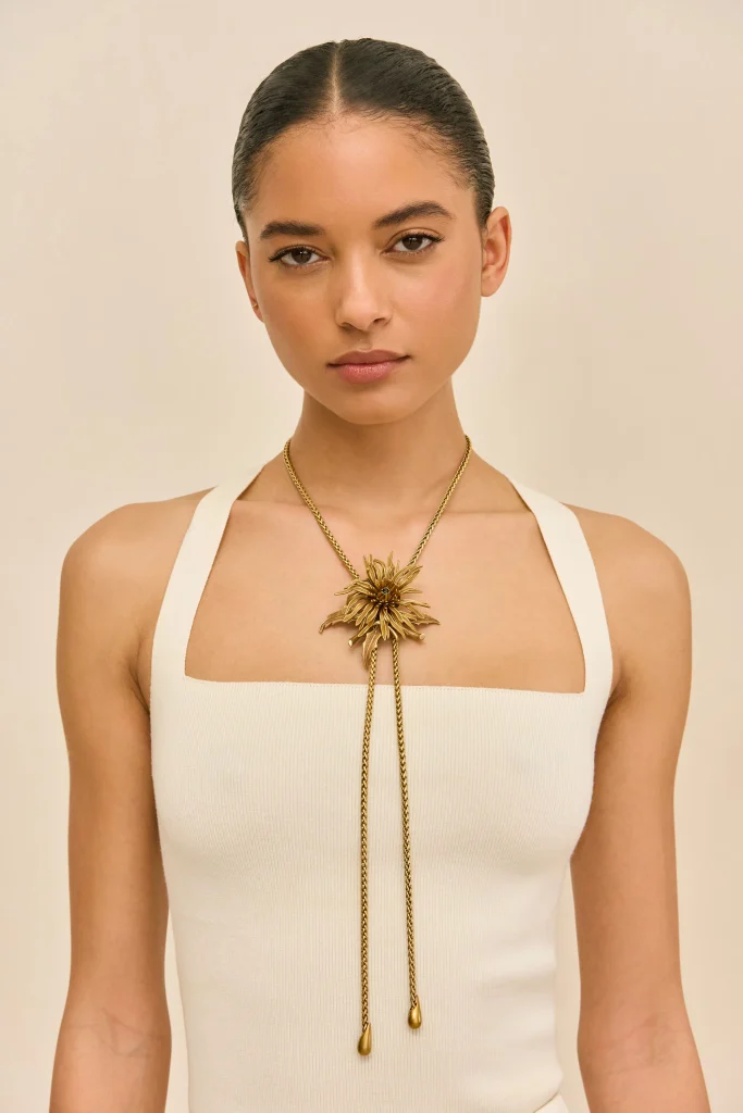 Woman wearing a white sleeveless top with a gold sunburst pendant necklace, facing the camera against a beige background.