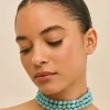 Close-up of a woman with eyes closed, wearing multiple turquoise beaded necklaces and a slicked-back hairstyle, head tilted to the side.