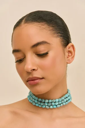 Close-up of a woman with eyes closed, wearing multiple turquoise beaded necklaces and a slicked-back hairstyle, head tilted to the side.