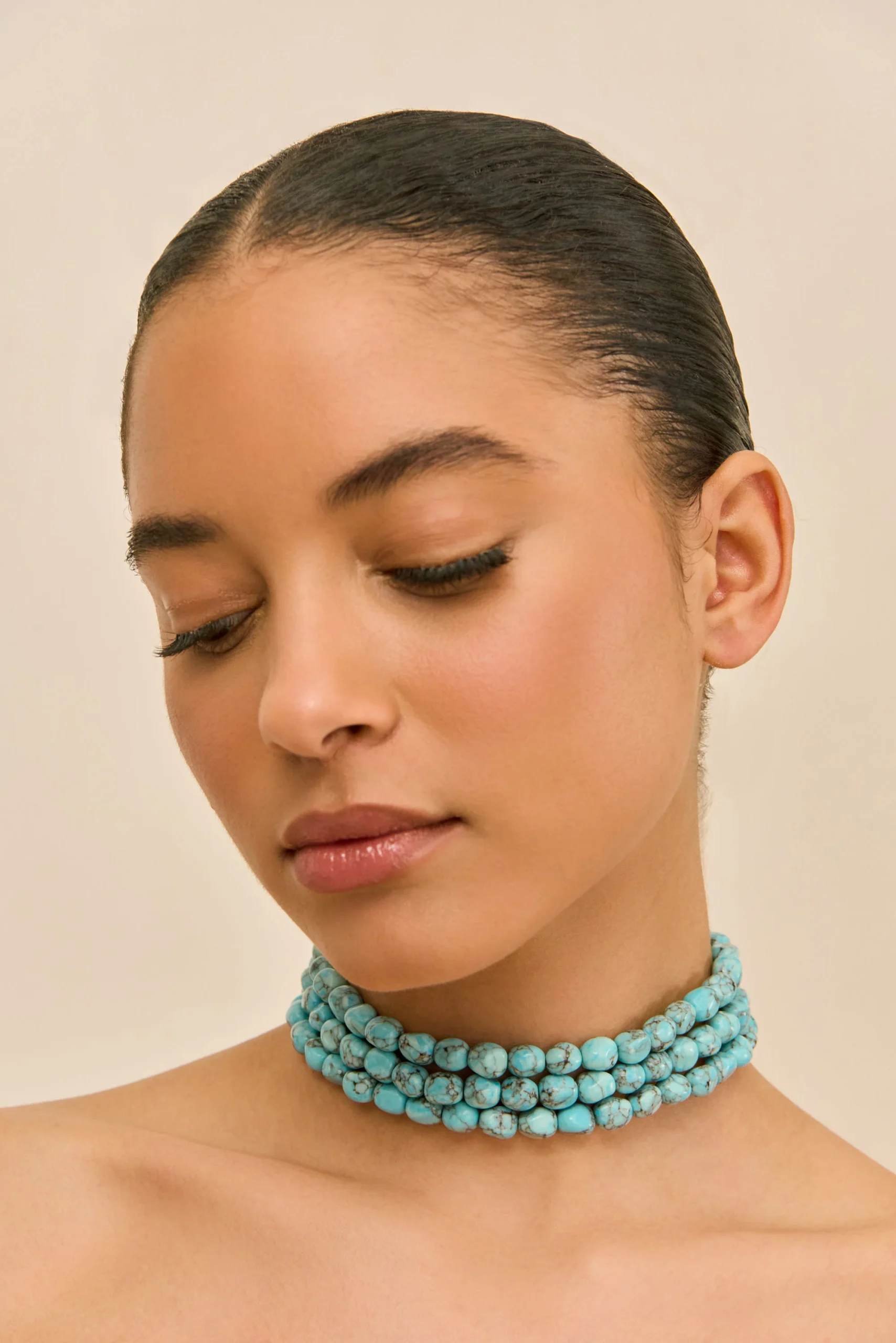 Close-up of a woman with eyes closed, wearing multiple turquoise beaded necklaces and a slicked-back hairstyle, head tilted to the side.