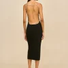 Woman standing with back to camera, wearing a backless black dress with pearl straps and black heeled sandals against a beige background