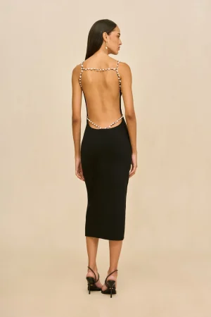 Woman standing with back to camera, wearing a backless black dress with pearl straps and black heeled sandals against a beige background