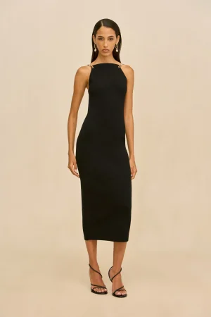 Woman in a black halter-neck midi dress with gold drop earrings, standing against a neutral beige backdrop.