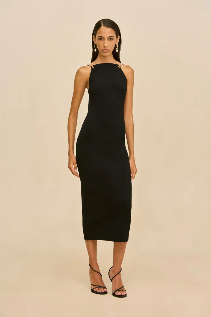 Woman in a black halter-neck midi dress with gold drop earrings, standing against a neutral beige backdrop.