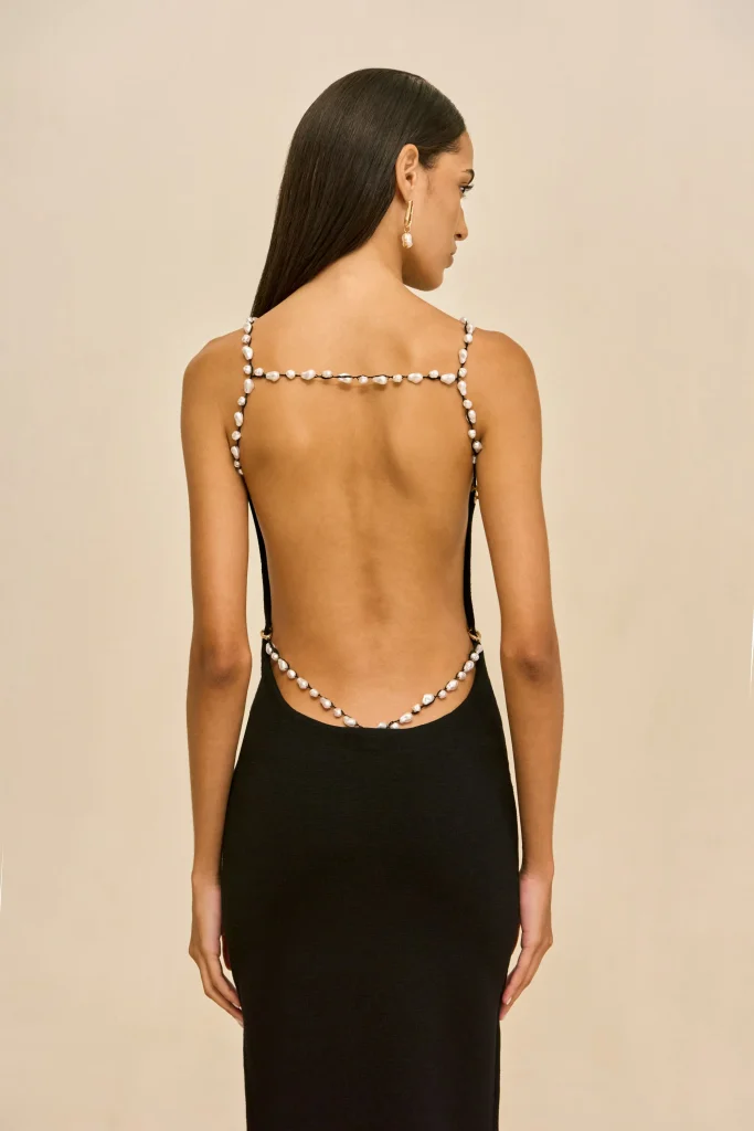 Back view of woman in a black backless dress wearing a pearl strap and gold hoop earrings against a beige background.