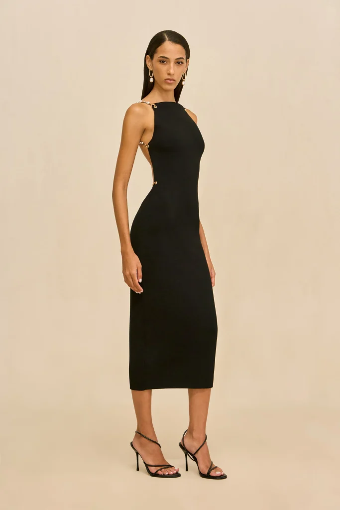 Woman in a fitted black midi dress with gold-tone shoulder details, standing against a neutral background, wearing strappy black heels and drop earrings.