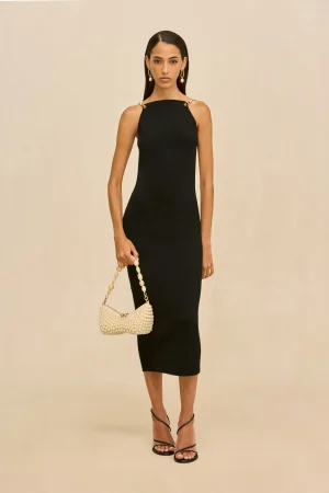 Full-length view of a woman in a black sleeveless dress, holding a pearl-bead handbag and wearing pearl earrings against a beige background.