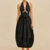 Full-length view of a woman in a black halter dress with waist cutouts and a gathered skirt, wearing black sandals against a beige backdrop.