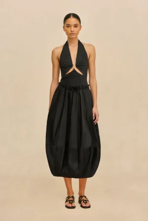 Full-length view of a woman in a black halter dress with waist cutouts and a gathered skirt, wearing black sandals against a beige backdrop.