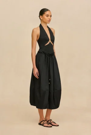Model wearing a black halter-neck dress with side cut-outs and a gathered, flowing skirt, standing in a beige studio background and wearing black flat sandals.