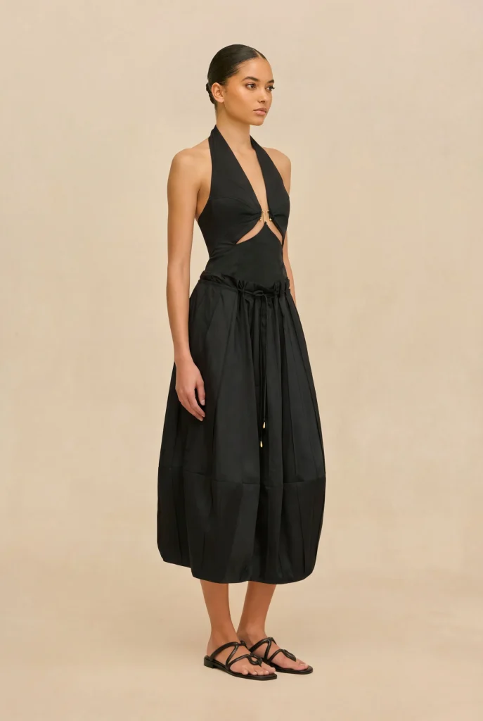 Model wearing a black halter-neck dress with side cut-outs and a gathered, flowing skirt, standing in a beige studio background and wearing black flat sandals.