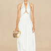 Model wearing a white halter dress with a front cutout, holding a gold woven handbag against a beige backdrop.
