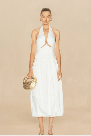 Model wearing a white halter dress with a front cutout, holding a gold woven handbag against a beige backdrop.
