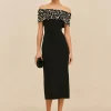 Slender woman wearing an off-shoulder black evening gown with a beaded, pearl-studded bodice, holding a small black clutch.