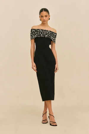 Model wearing a black off-shoulder evening dress with a jeweled, beaded bodice, standing against a neutral beige background.