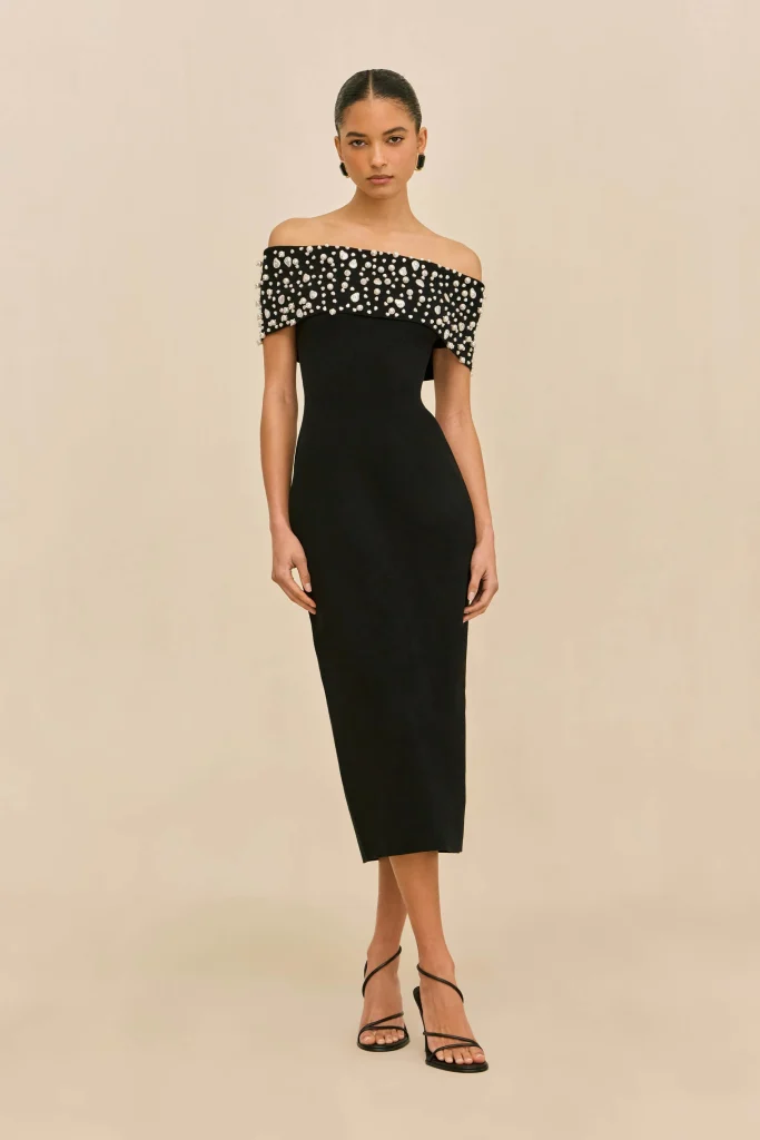 Model wearing a black off-shoulder evening dress with a jeweled, beaded bodice, standing against a neutral beige background.
