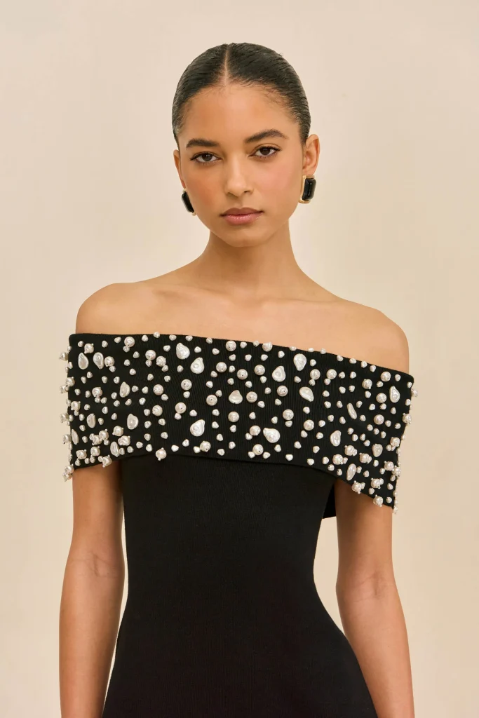 Portrait of a woman with slicked-back hair wearing an off-the-shoulder black dress embellished with white pearls, posing against a beige backdrop.
