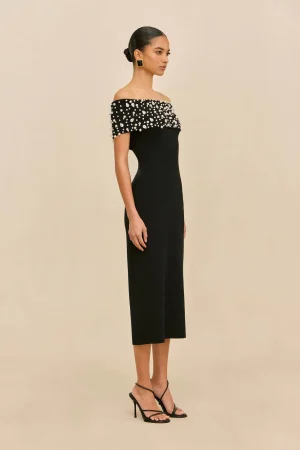 Model in a black off-the-shoulder dress with pearl embellishments, square earrings, and strappy black heels, shown in profile.