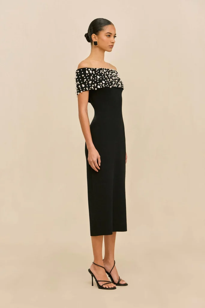 Model in a black off-the-shoulder dress with pearl embellishments, square earrings, and strappy black heels, shown in profile.