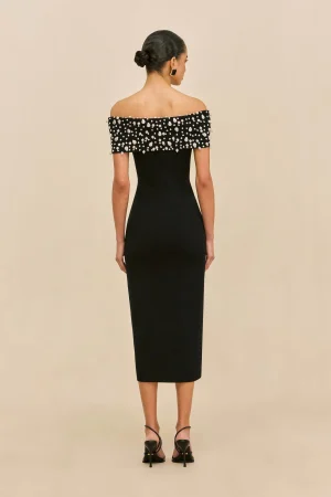 Back view of a woman in a black off-the-shoulder evening dress with a beaded, pearl-studded neckline, her hair pulled into a neat bun, standing against a beige background, wearing black sandals.