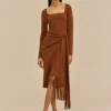 Model wearing a rust brown square‑neck wrap dress with a waist tie and fringe hem, paired with gold hoop earrings and tan sandals.