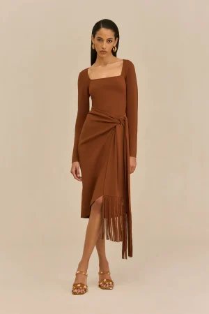 Model wearing a rust brown square‑neck wrap dress with a waist tie and fringe hem, paired with gold hoop earrings and tan sandals.