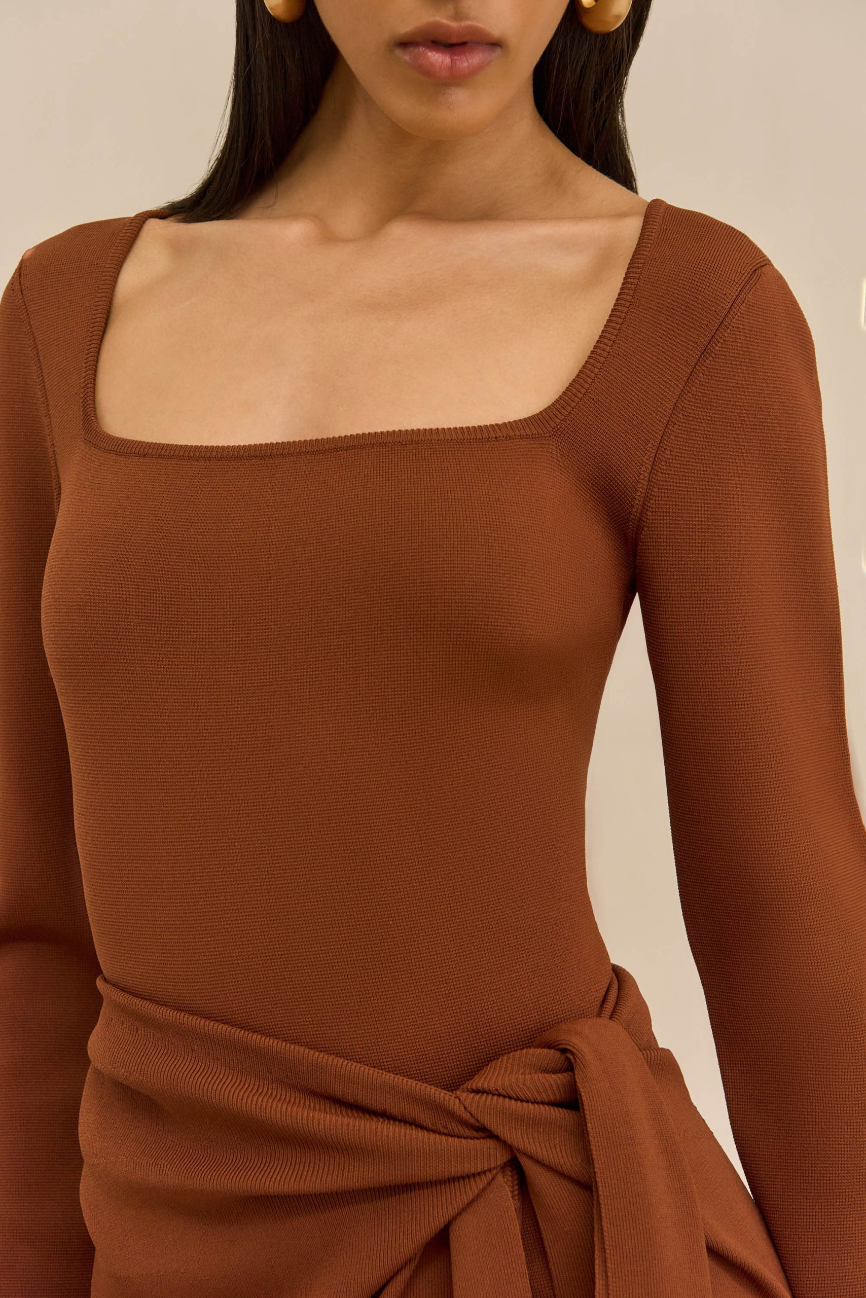 Close-up of a person wearing a rust brown ribbed knit top with a square neckline and a tied waist belt.