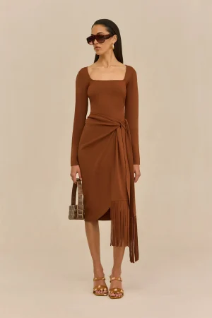 Woman in a brown wrap dress with long sleeves, tied at the waist, wearing oversized brown sunglasses and gold earrings, holding a square, checkered handbag, standing against a beige background.