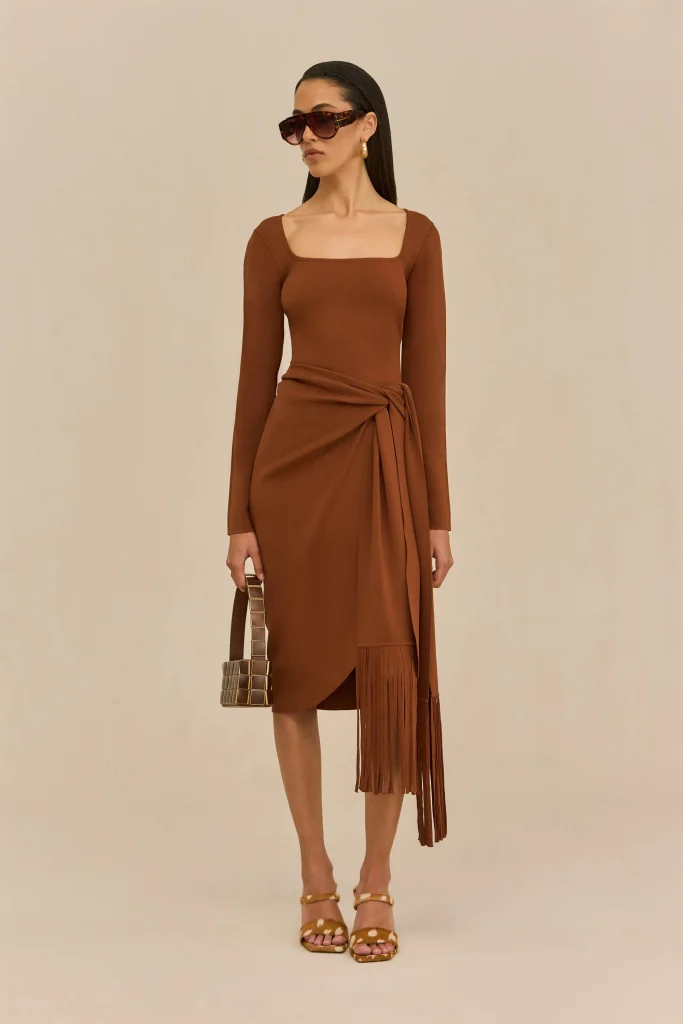 Woman in a brown wrap dress with long sleeves, tied at the waist, wearing oversized brown sunglasses and gold earrings, holding a square, checkered handbag, standing against a beige background.