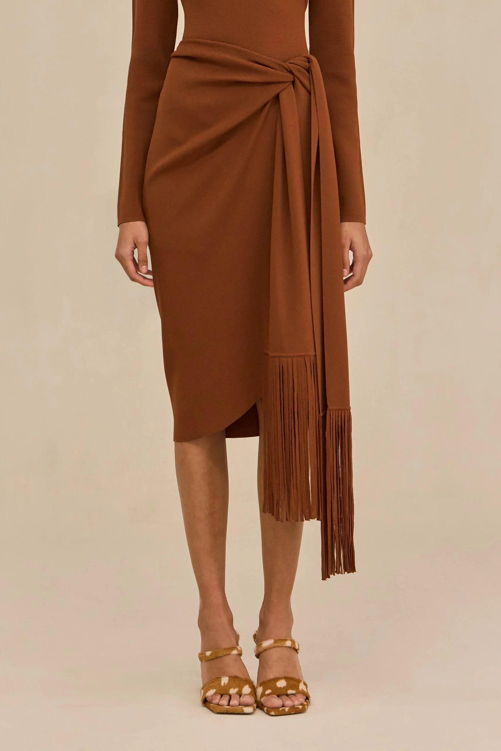 Rust-brown wrap dress with a tied waist and long fringe tassels, paired with tan sandals.