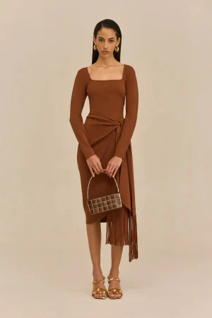 Full-body shot of a woman in a brown wrap dress with fringe, holding a small square handbag, against a beige background.