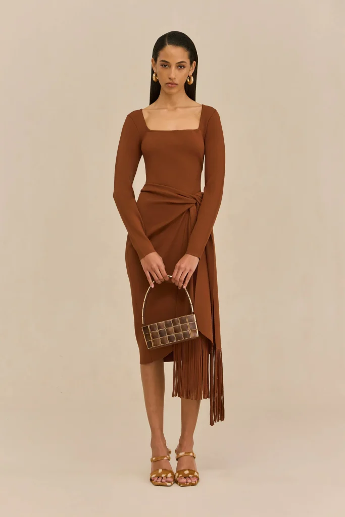 Full-body shot of a woman in a brown wrap dress with fringe, holding a small square handbag, against a beige background.