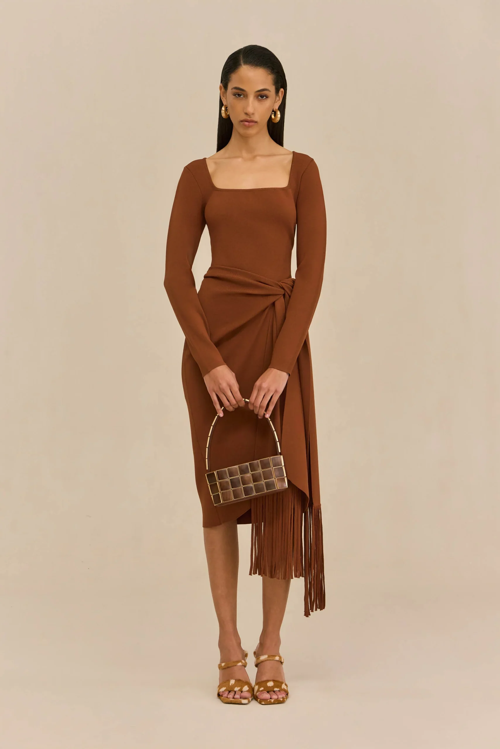 Full-body shot of a woman in a brown wrap dress with fringe, holding a small square handbag, against a beige background.