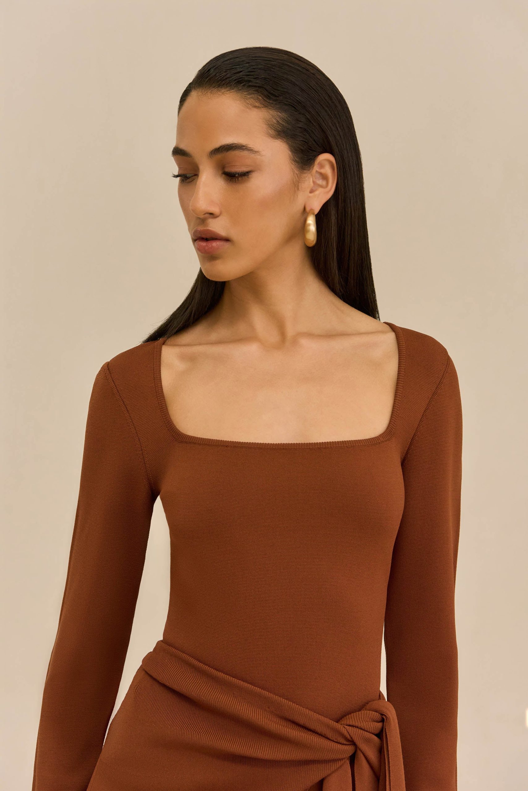 Woman with sleek dark hair and a rust-colored square-neck dress, wearing a gold hoop earring, looking downward against a neutral background.