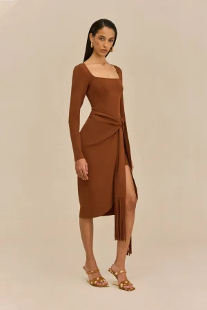 Woman standing in a brown wrap dress with a tied sash, wearing gold hoop earrings and tan sandals against a beige backdrop.