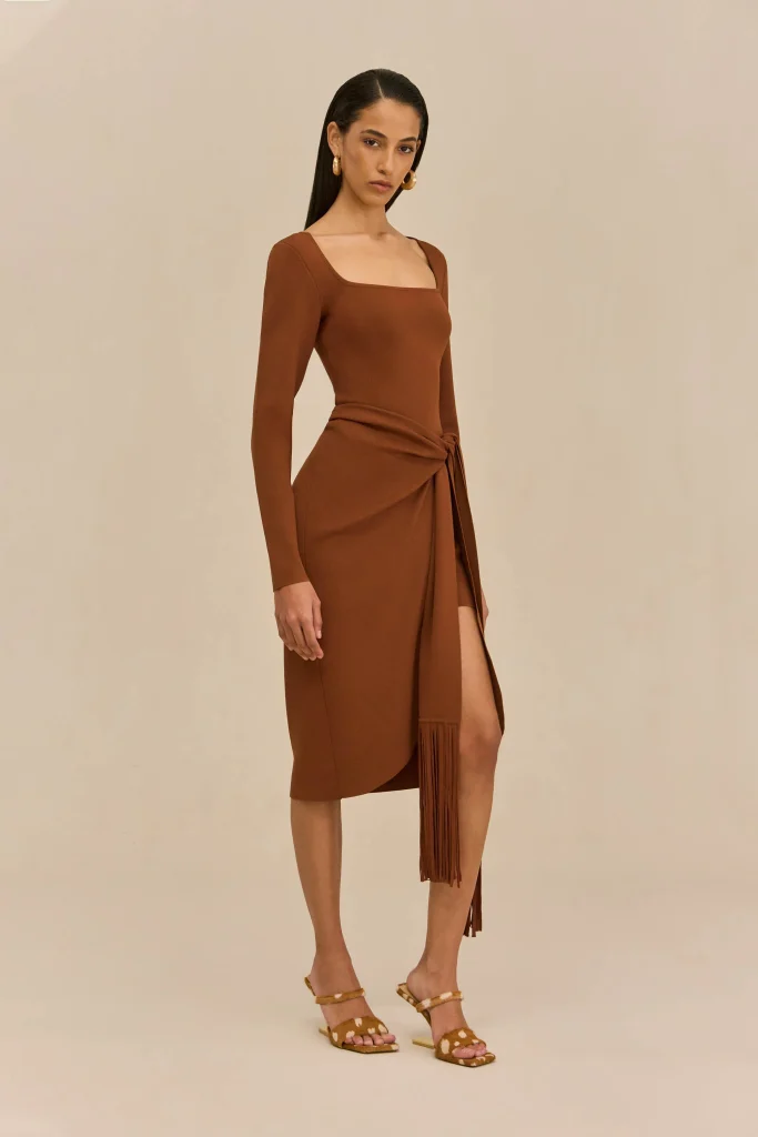 Woman standing in a brown wrap dress with a tied sash, wearing gold hoop earrings and tan sandals against a beige backdrop.