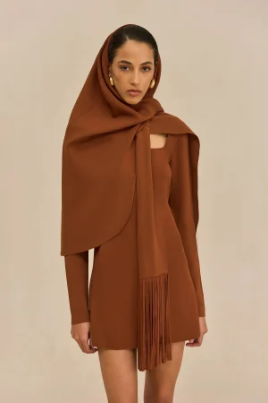 Woman wearing a rust-brown hijab and matching dress, standing against a light beige background with a calm expression.