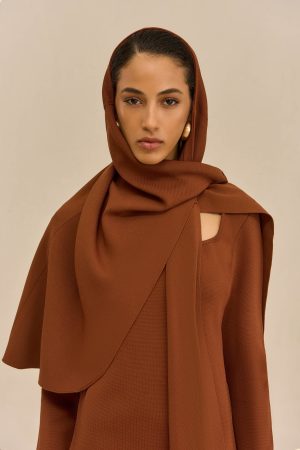 Close-up of a woman wearing a brown hijab, facing the camera with a neutral beige background, gold hoop earring visible on one side.