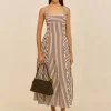 Woman modeling a brown-and-white striped sleeveless maxi dress with square neckline, gold hoop earrings, clear sandals, and a dark handbag against a neutral backdrop.