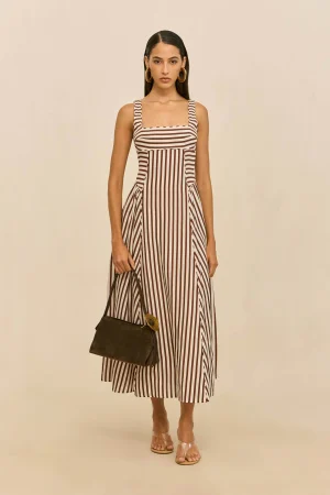Woman modeling a brown-and-white striped sleeveless maxi dress with square neckline, gold hoop earrings, clear sandals, and a dark handbag against a neutral backdrop.