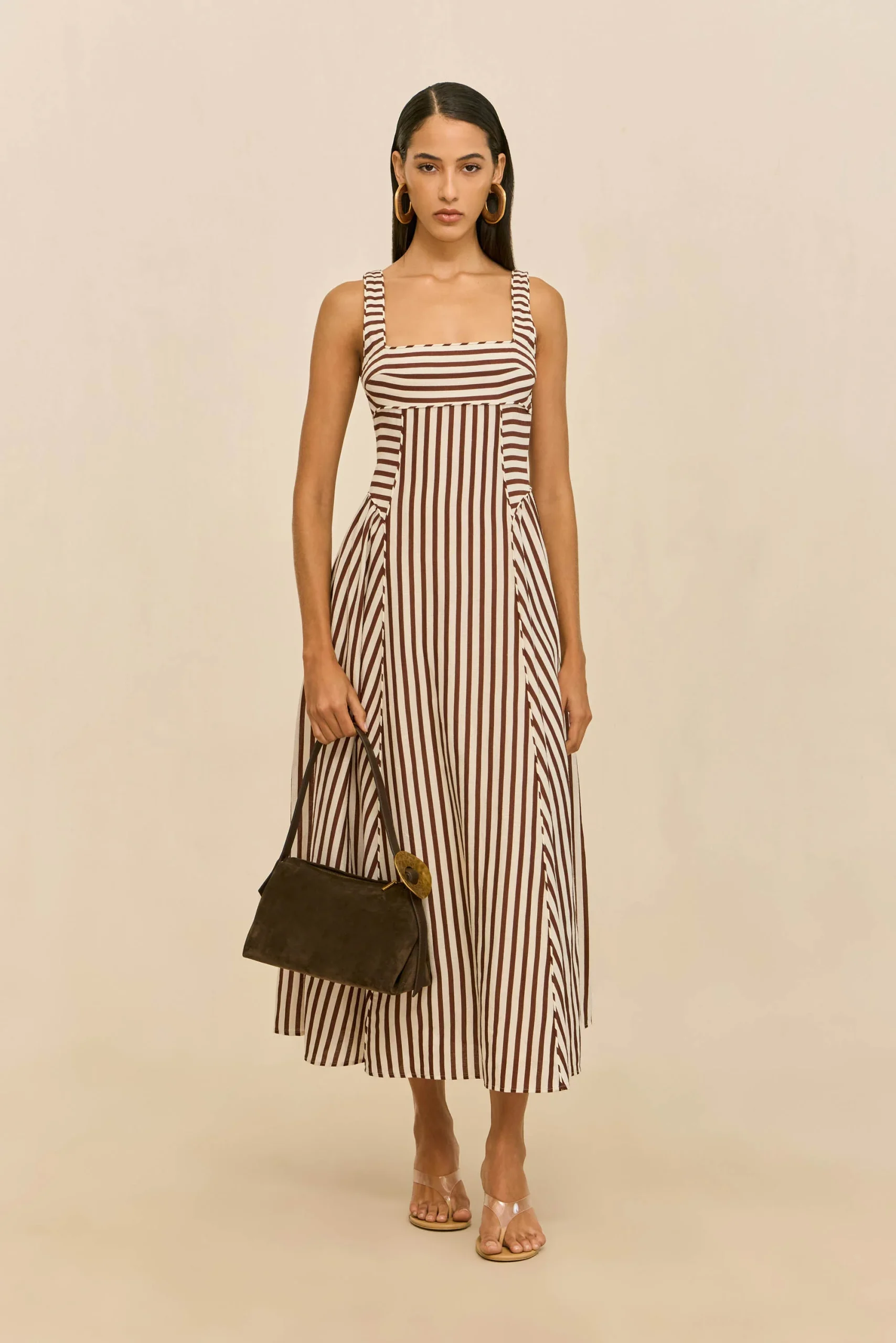 Woman modeling a brown-and-white striped sleeveless maxi dress with square neckline, gold hoop earrings, clear sandals, and a dark handbag against a neutral backdrop.