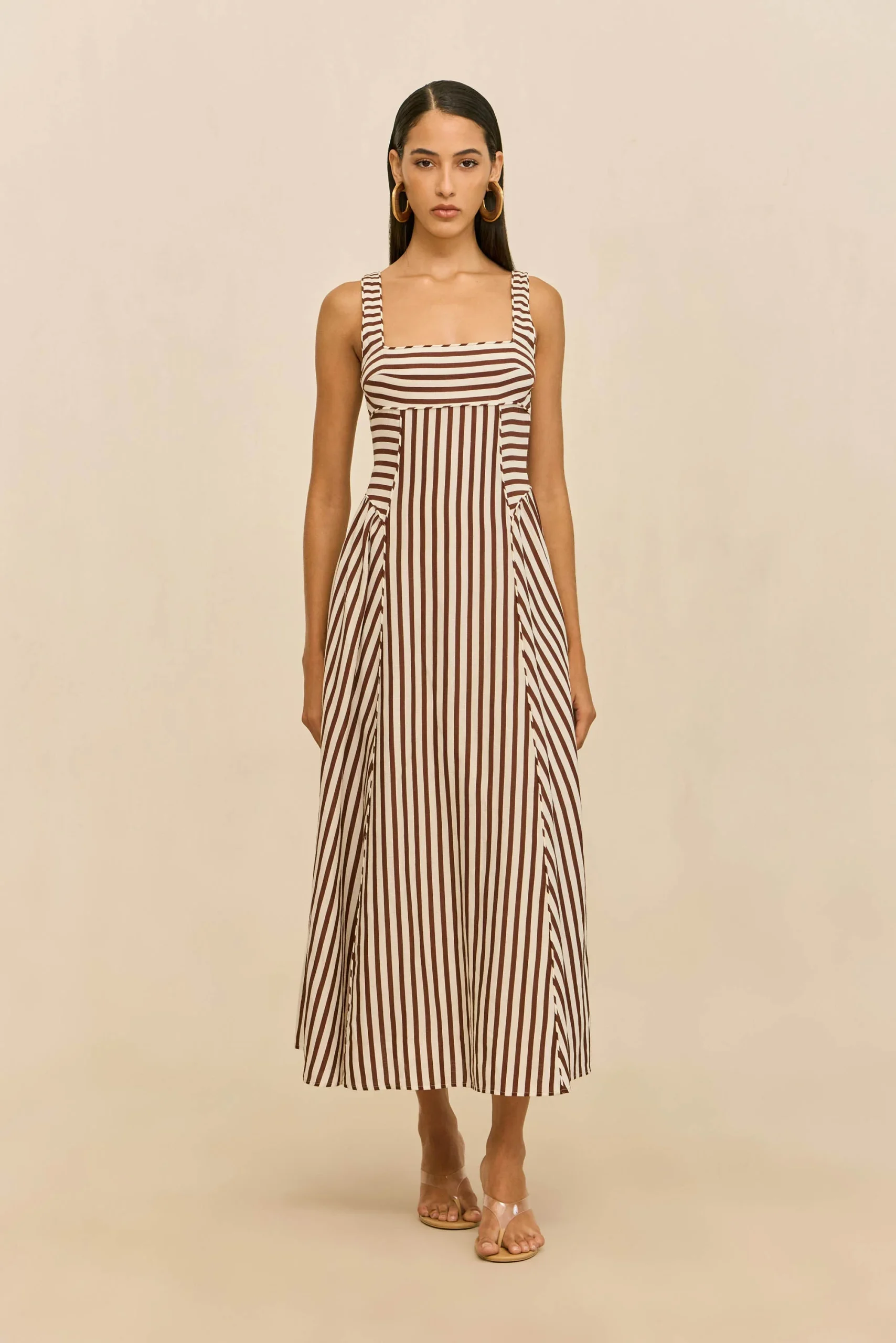 Full-length model wearing a brown-and-white vertical striped maxi dress with a squared neckline and large gold hoop earrings, standing against a beige backdrop.