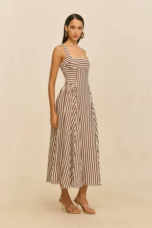 Model wearing a brown and white vertical-striped sleeveless maxi dress with sandals against a beige backdrop, posing sideways.