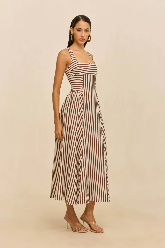 Model wearing a brown and white vertical-striped sleeveless maxi dress with sandals against a beige backdrop, posing sideways.