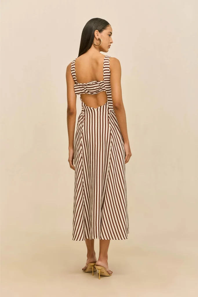 Woman in a brown-and-white striped backless dress with a bow, viewed from behind, wearing gold hoop earrings.