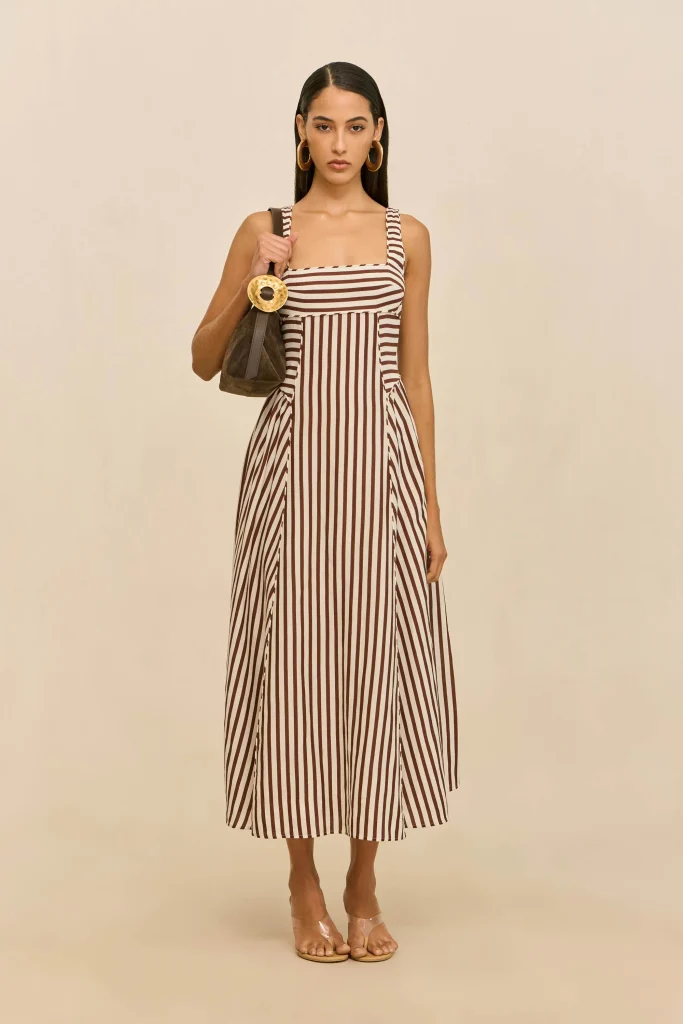 Full-body shot of a woman in a vertical brown-and-white striped maxi dress, holding a brown handbag with a large circular gold handle, standing against a neutral beige background.