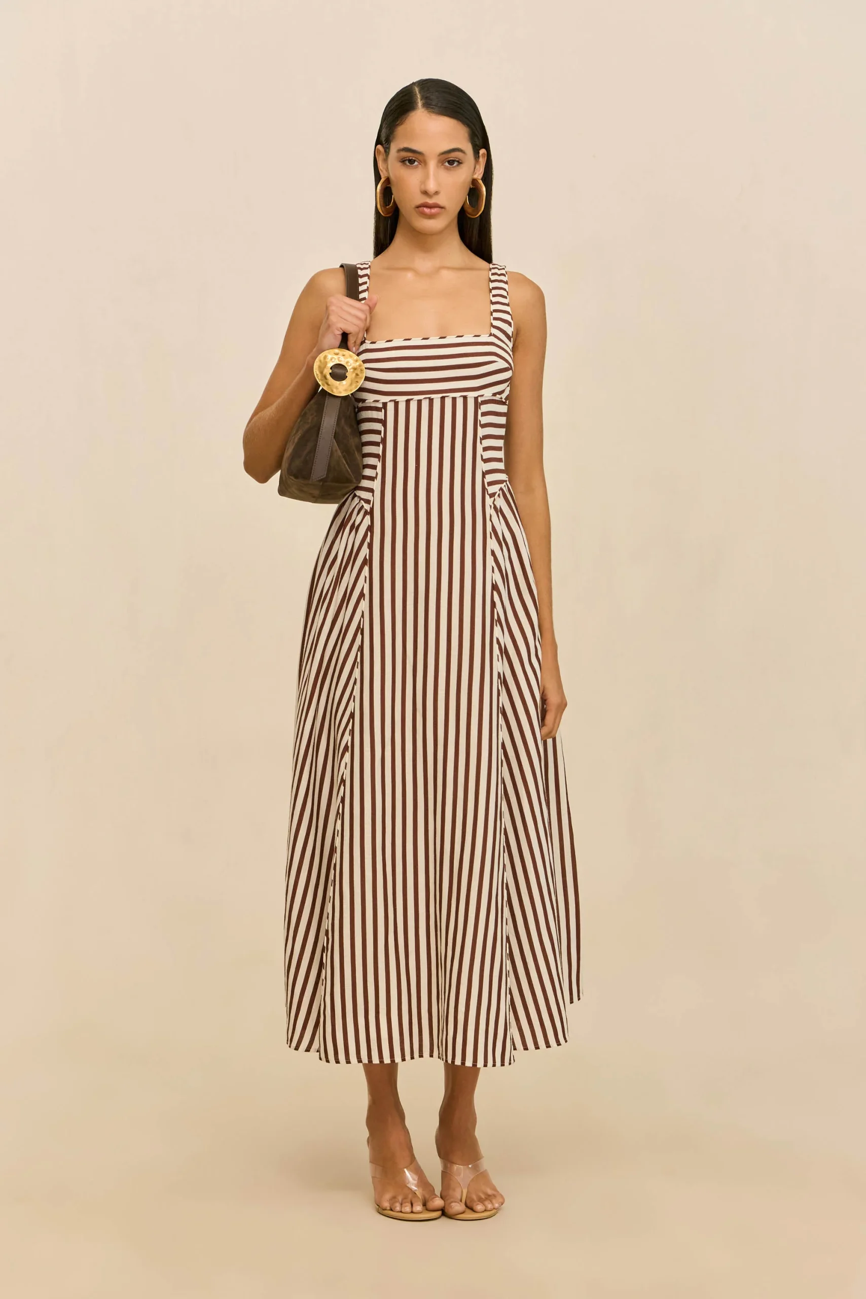 Full-body shot of a woman in a vertical brown-and-white striped maxi dress, holding a brown handbag with a large circular gold handle, standing against a neutral beige background.
