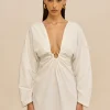 Woman wearing a white, plunging-neckline dress with a ruched center knot and gold hoop earrings, standing against a beige background.