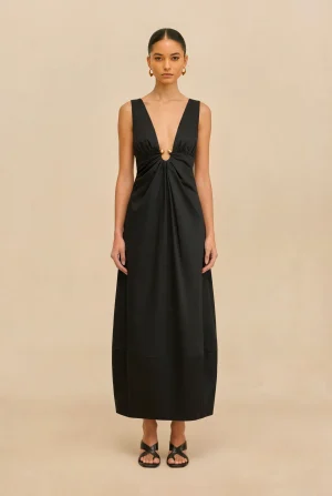 Woman standing in a black sleeveless dress with a deep V neckline and gold accents, against a beige backdrop, wearing open-toed sandals.
